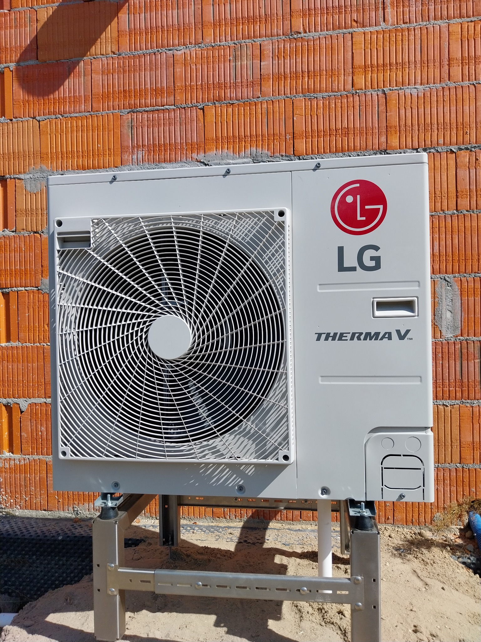 Zewnętrzna jednostka pompy ciepła LG Therma V zamontowana na metalowej konstrukcji przy ścianie z cegły ceramicznej, widoczne przyłącza i cień na murze.