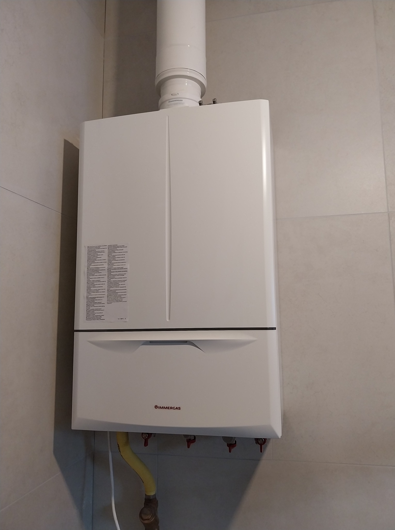 Biały, wiszący piec gazowy Immergas z żółtym przewodem gazowym na tle jasnych, kwadratowych płytek. Widoczne zawory i rura odprowadzająca spaliny.