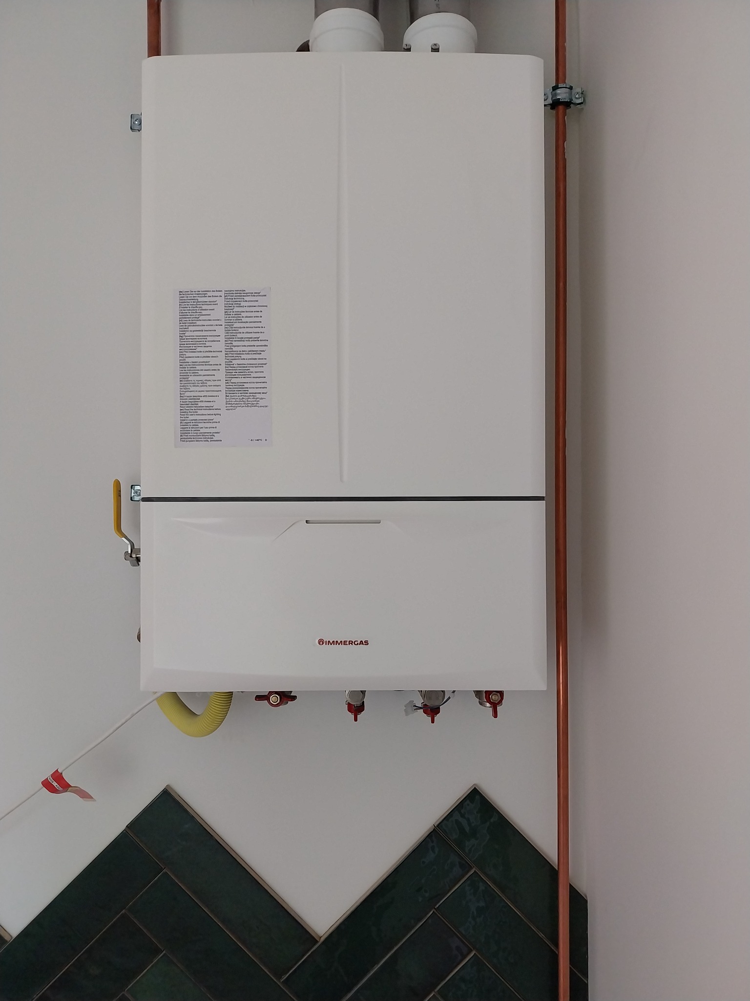 Biały, wiszący piec gazowy Immergas z miedzianymi rurami na tle białej ściany z dekoracyjnymi, ciemnozielonymi płytkami ułożonymi w jodełkę.