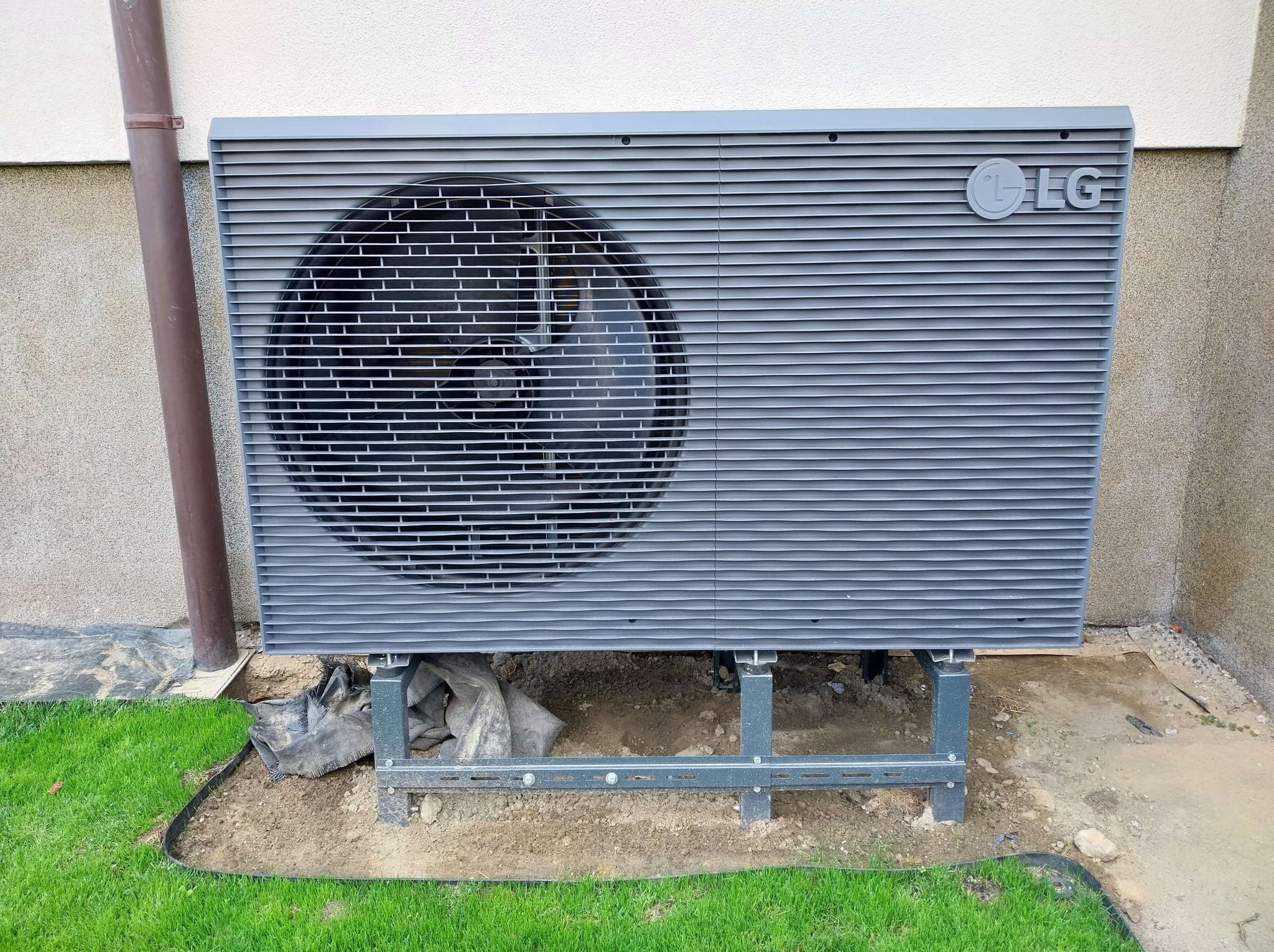 Szara pompa ciepła LG na metalowej konstrukcji, zainstalowana przy ścianie budynku z fragmentem trawnika. Widoczne elementy montażowe i logo producenta.