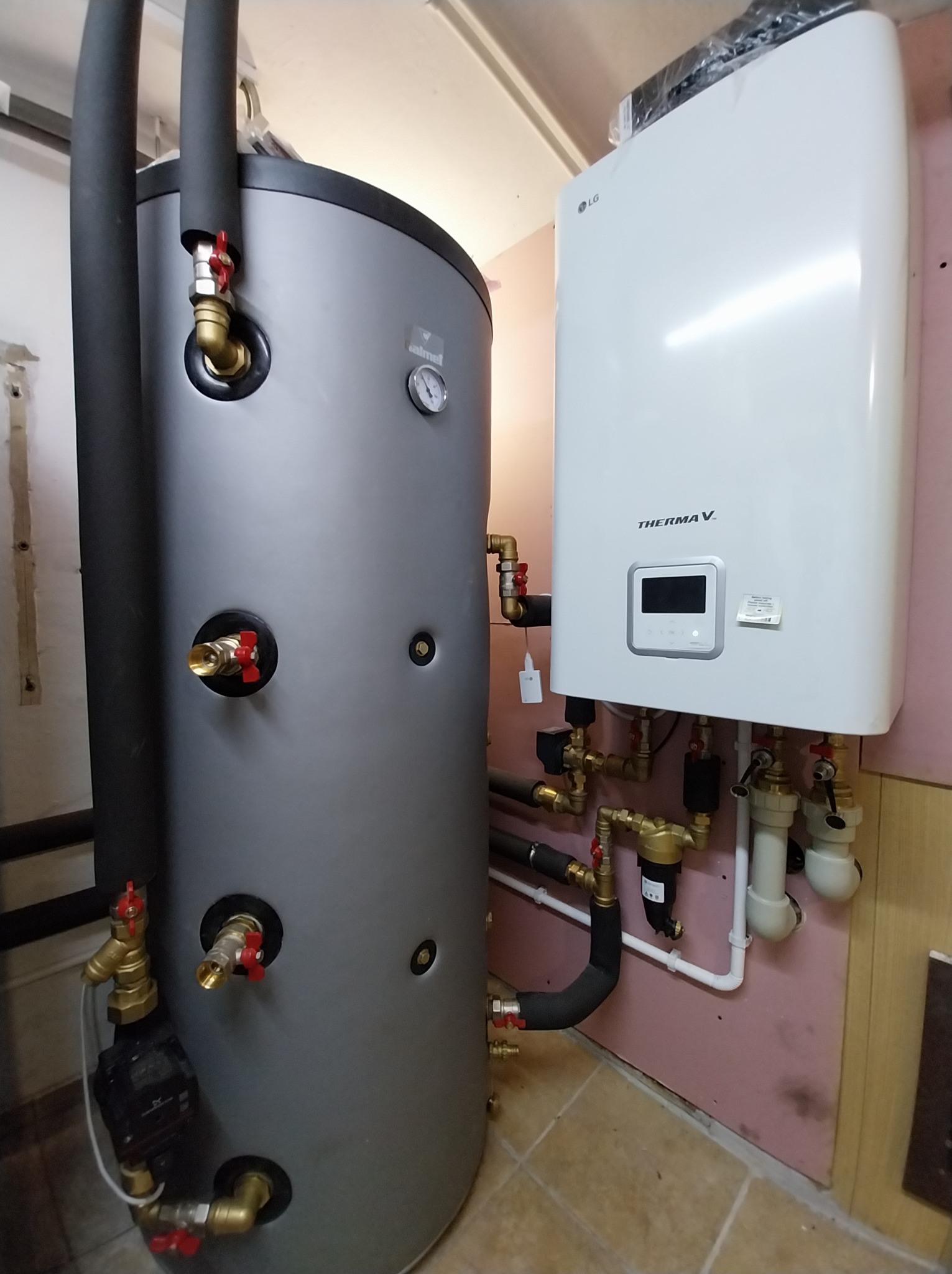 Szary zbiornik akumulacyjny ciepła z czarną izolacją rur i biała pompa ciepła LG Therma V na różowej ścianie, widoczne liczne zawory i rury.
