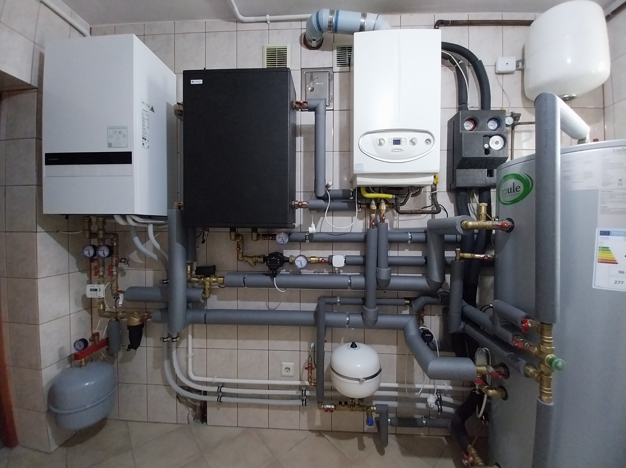 Złożona instalacja grzewcza w kotłowni: dwa piece (biały i czarny), zbiornik, liczne rury izolowane szarą pianką, manometry i zawory na tle płytek ceramicznych.