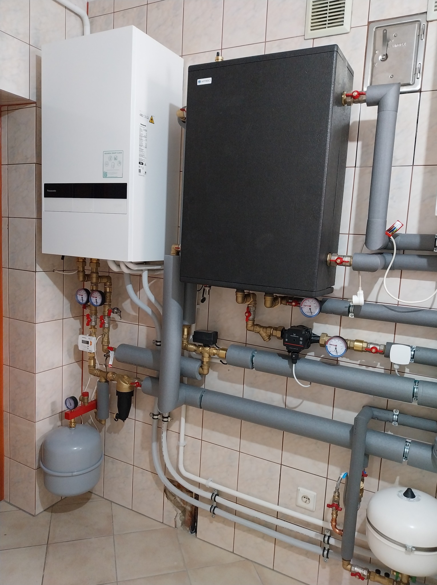 Złożona instalacja grzewcza z białym piecem gazowym Panasonic i czarnym zasobnikiem, rury w szarej izolacji, manometry, zawory, na tle beżowych płytek.