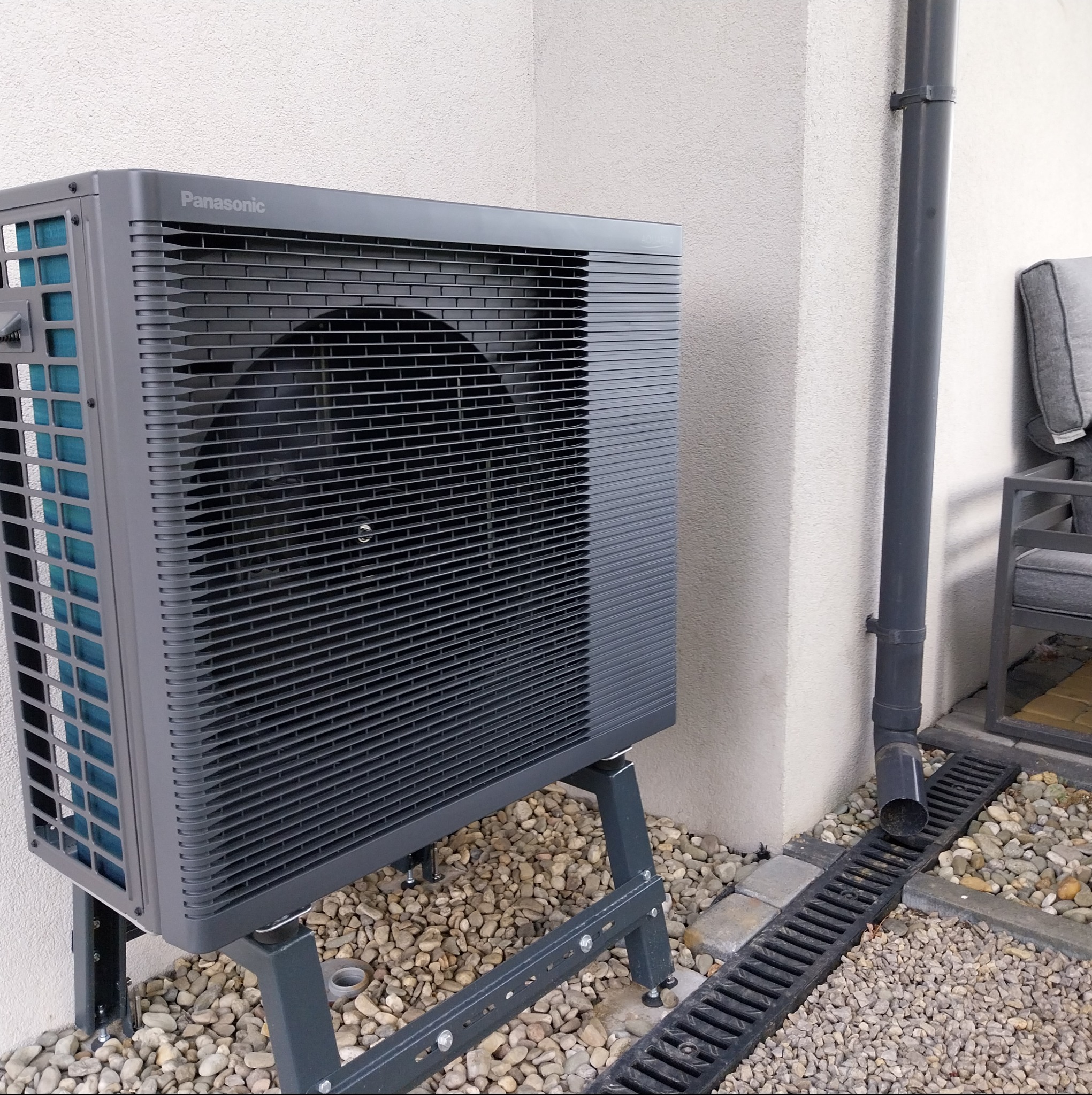 Szara pompa ciepła Panasonic na zewnątrz budynku, zamontowana na metalowej konstrukcji, z widoczną rurą spustową i kamiennym podłożem. Ujęcie z boku, w dziennym świetle.