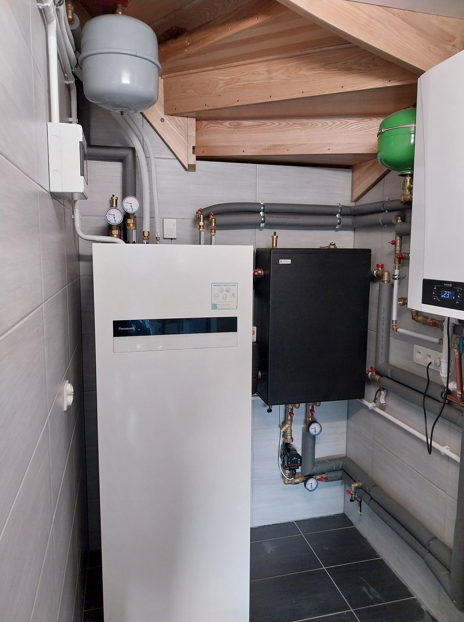 Nowoczesna instalacja grzewcza z pompą ciepła Panasonic Aquarea, zbiornikiem buforowym i osprzętem, w pomieszczeniu wykończonym szarą płytką. Widoczne rury i manometry.