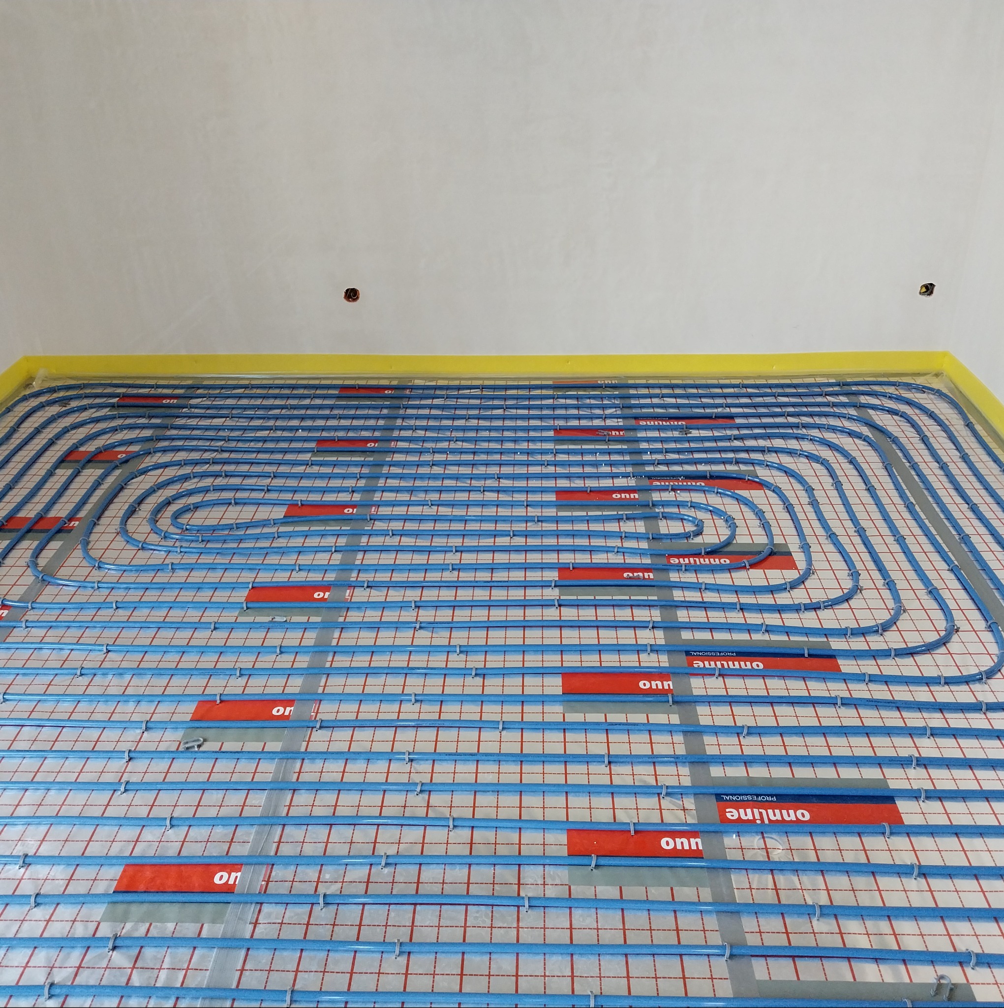 Niebieskie rury ogrzewania podłogowego ułożone na folii z czerwoną siatką, przymocowane plastikowymi uchwytami. Widok z góry, żółta taśma na ścianie.