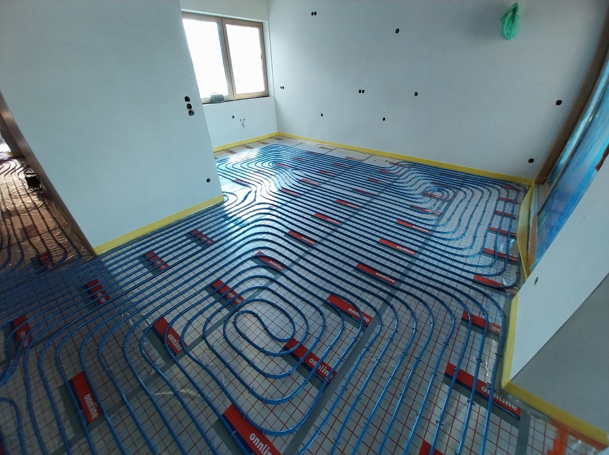 Niebieskie rury ogrzewania podłogowego ułożone spiralnie na folii izolacyjnej w jasnym pomieszczeniu z oknem. Widoczne czerwone elementy mocujące rury.