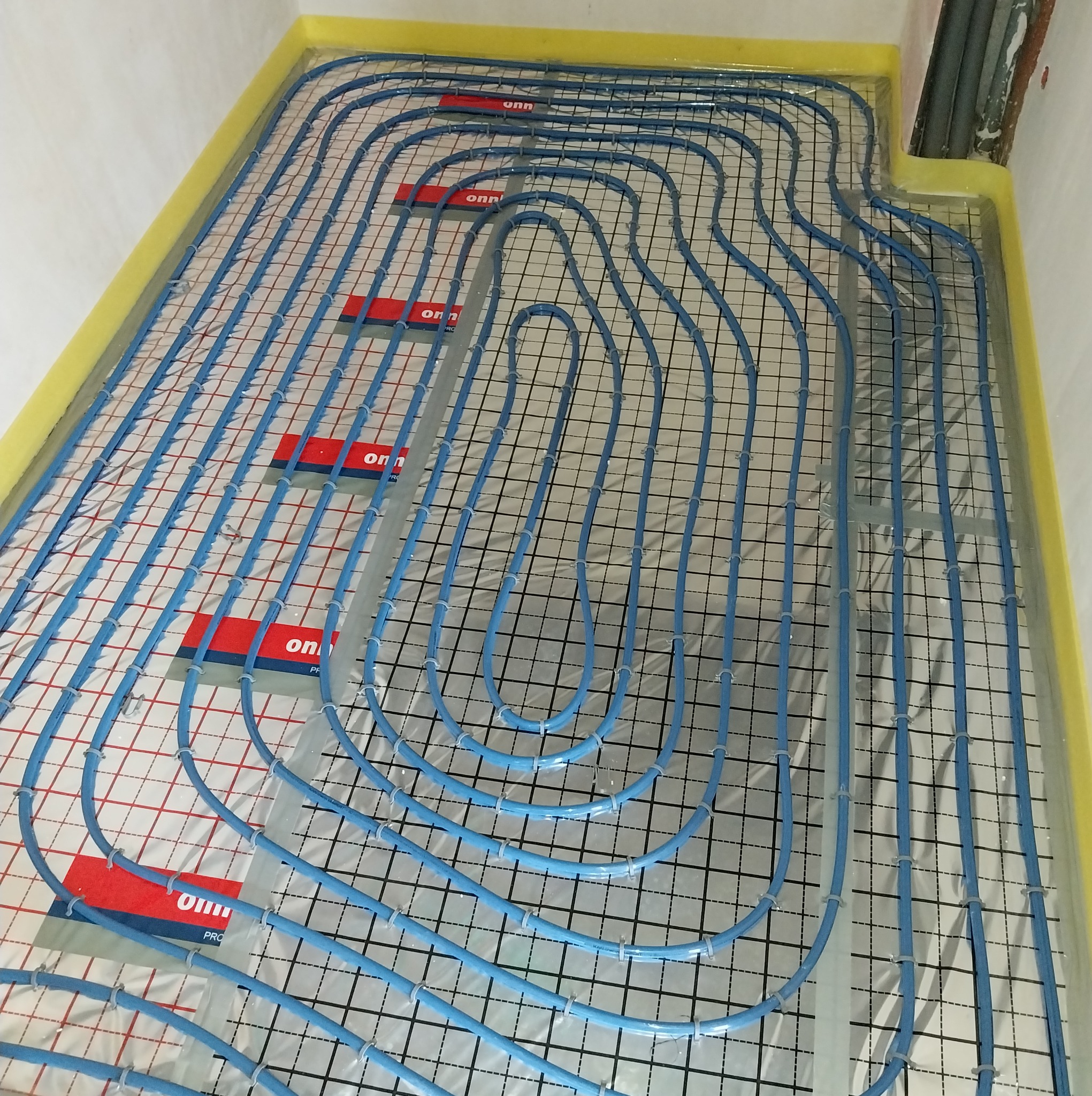 Niebieskie rury ogrzewania podłogowego ułożone na folii z siatką, przymocowane plastikowymi uchwytami. Żółta taśma izolacyjna wzdłuż ścian.