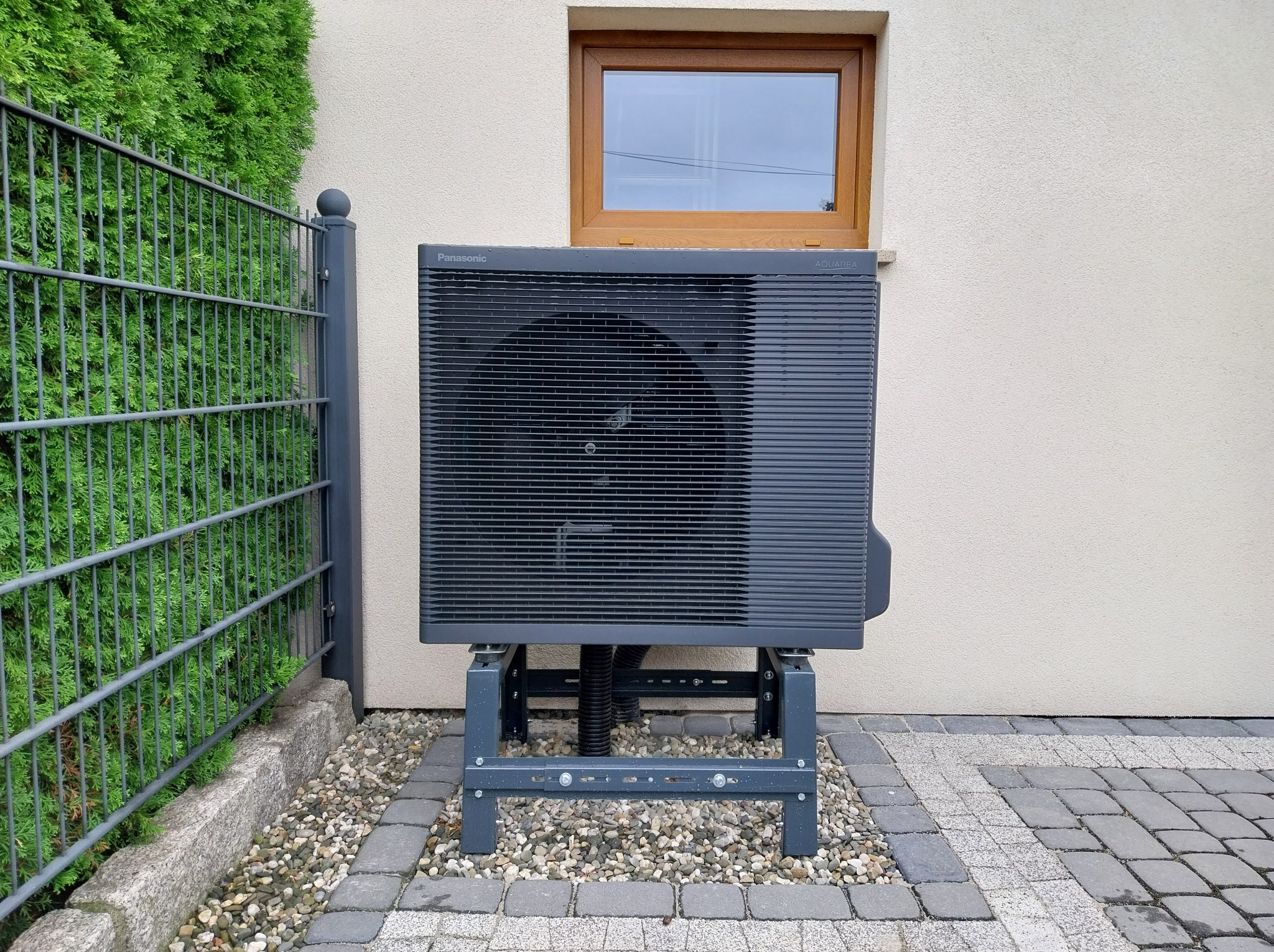 Szara pompa ciepła Panasonic Aquarea zamontowana na zewnątrz budynku, widoczna z boku, z zielonym żywopłotem w tle i oknem na ścianie.