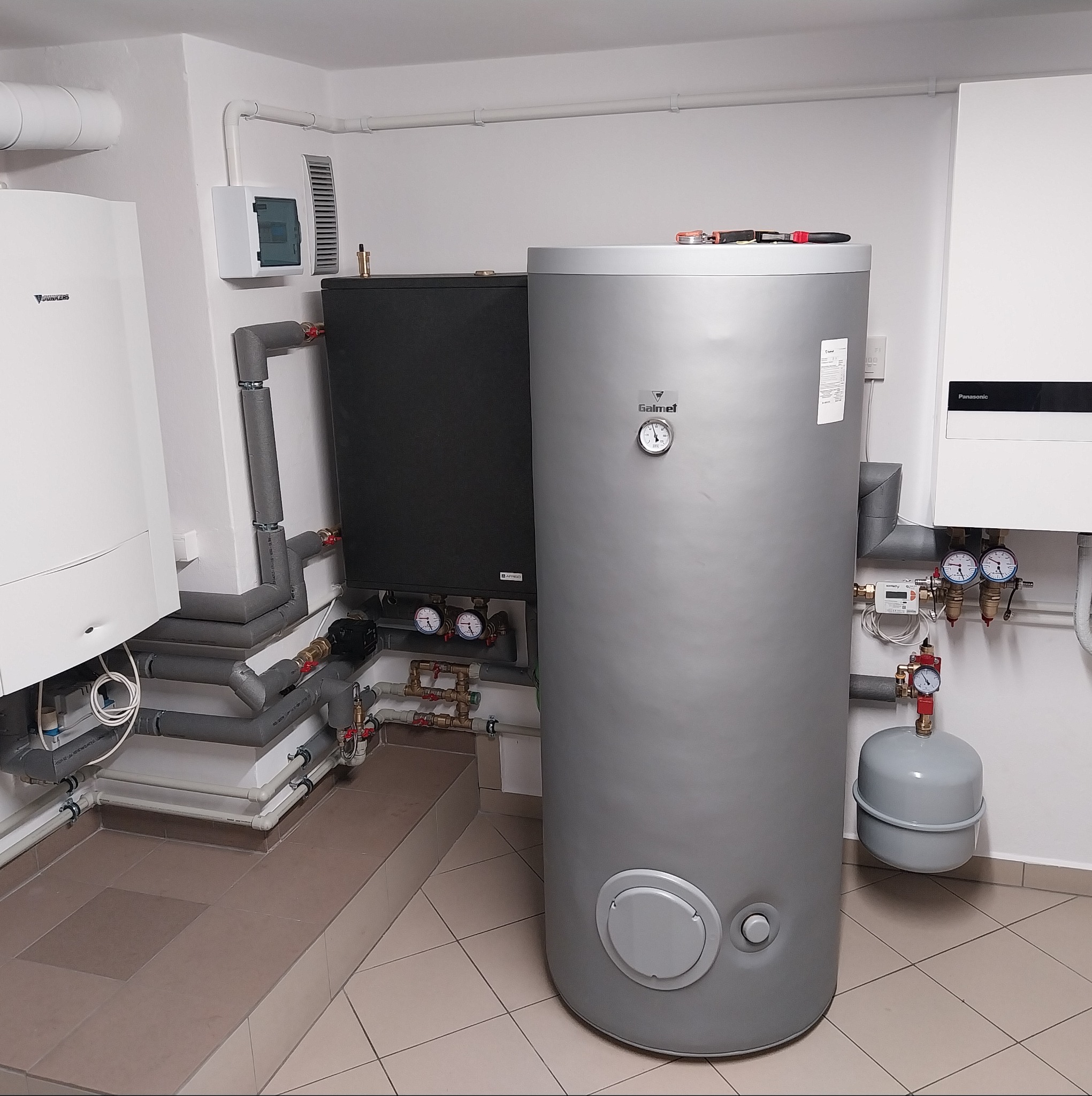Zestaw urządzeń grzewczych: piec gazowy Junkers, zasobnik Galmet i pompa ciepła Panasonic, połączone rurami w Siemianowicach Śląskich. Nowoczesna kotłownia.