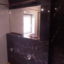 Wykończona łazienka z ciemnymi płytkami imitującymi marmur z wyraźnym użyleniem, chromowana armatura, duże lustro i drewnopodobna podłoga.