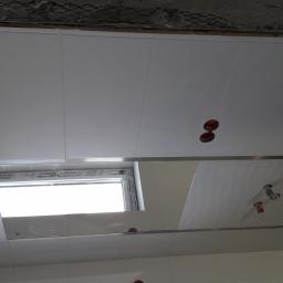 WIBERO Firma Budowlana - Biały sufit z paneli PCV z oknem i otworami na oświetlenie, widoczne niedokończone wykończenie sufitu nad panelami.