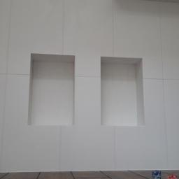 WIBERO Firma Budowlana - Ściana wykończona białymi płytami z dwoma wnękami, widoczne fugi i fragment drewnopodobnej podłogi.