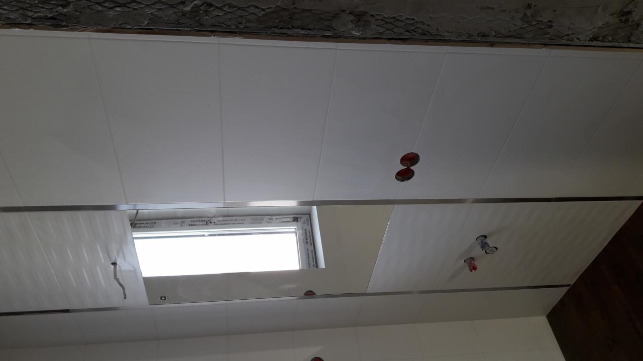 Biały sufit z paneli PCV z oknem i otworami na oświetlenie, widoczne niedokończone wykończenie sufitu nad panelami.