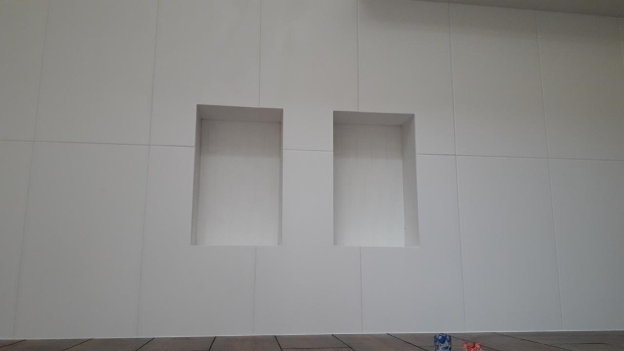 Ściana wykończona białymi płytami z dwoma wnękami, widoczne fugi i fragment drewnopodobnej podłogi.