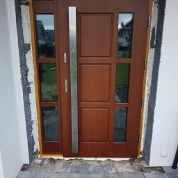 Świat Drzwi - Brązowe drzwi zewnętrzne z szybami i długim, srebrnym uchwytem, w trakcie montażu. Widoczne pianka montażowa i surowy mur. Ujęcie z bliska, z zewnątrz budynku.