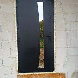 Świat Drzwi - Czarne, nowoczesne drzwi z szybą, osadzone w surowym murze z białych bloczków. Widoczna pianka montażowa i drewniane podpory. Ujęcie z bliska, z zewnątrz budynku.