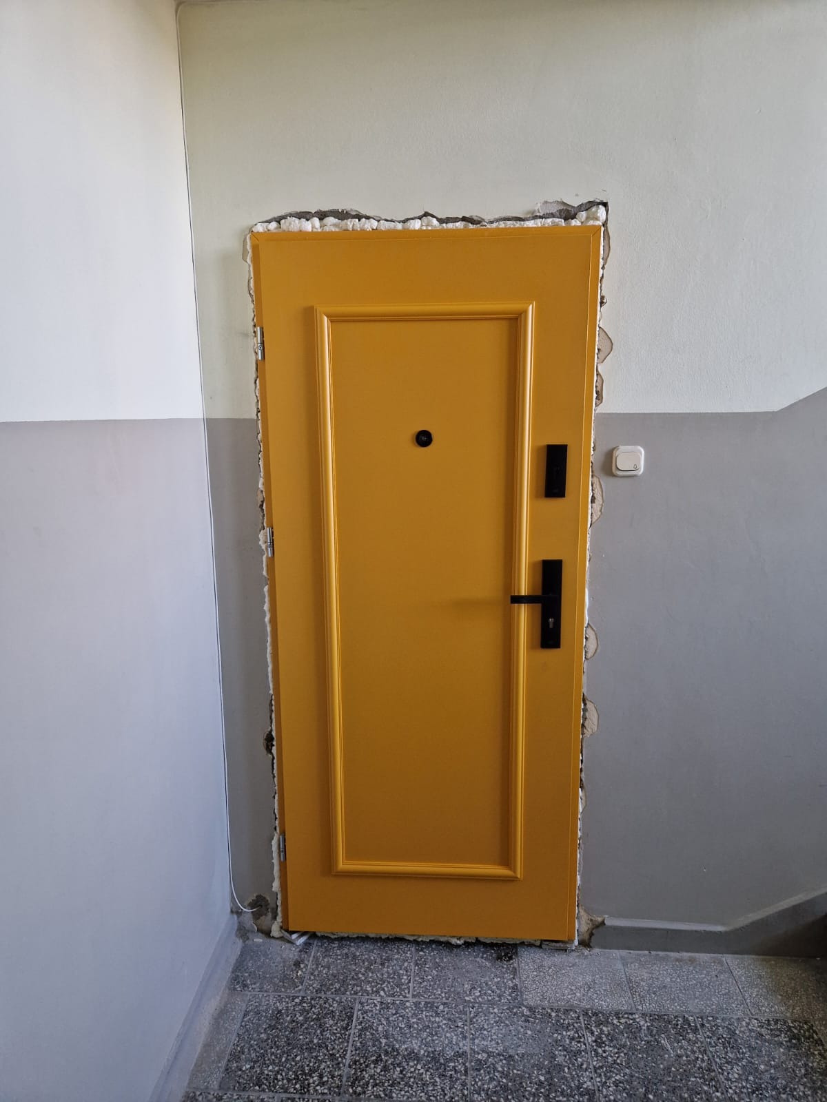Żółte drzwi z czarną klamką, osadzone w surowym otworze, widoczne ślady pianki montażowej. Ujęcie na klatce schodowej, z bliska, z wyraźnym detalem wykończenia.