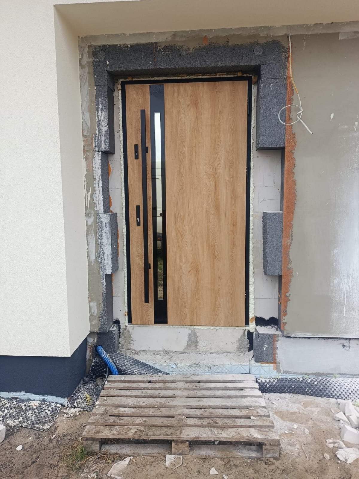 Montaż drzwi zewnętrznych z drewnopodobnym wykończeniem i czarnymi detalami, widoczne ocieplenie budynku w trakcie prac wykończeniowych.