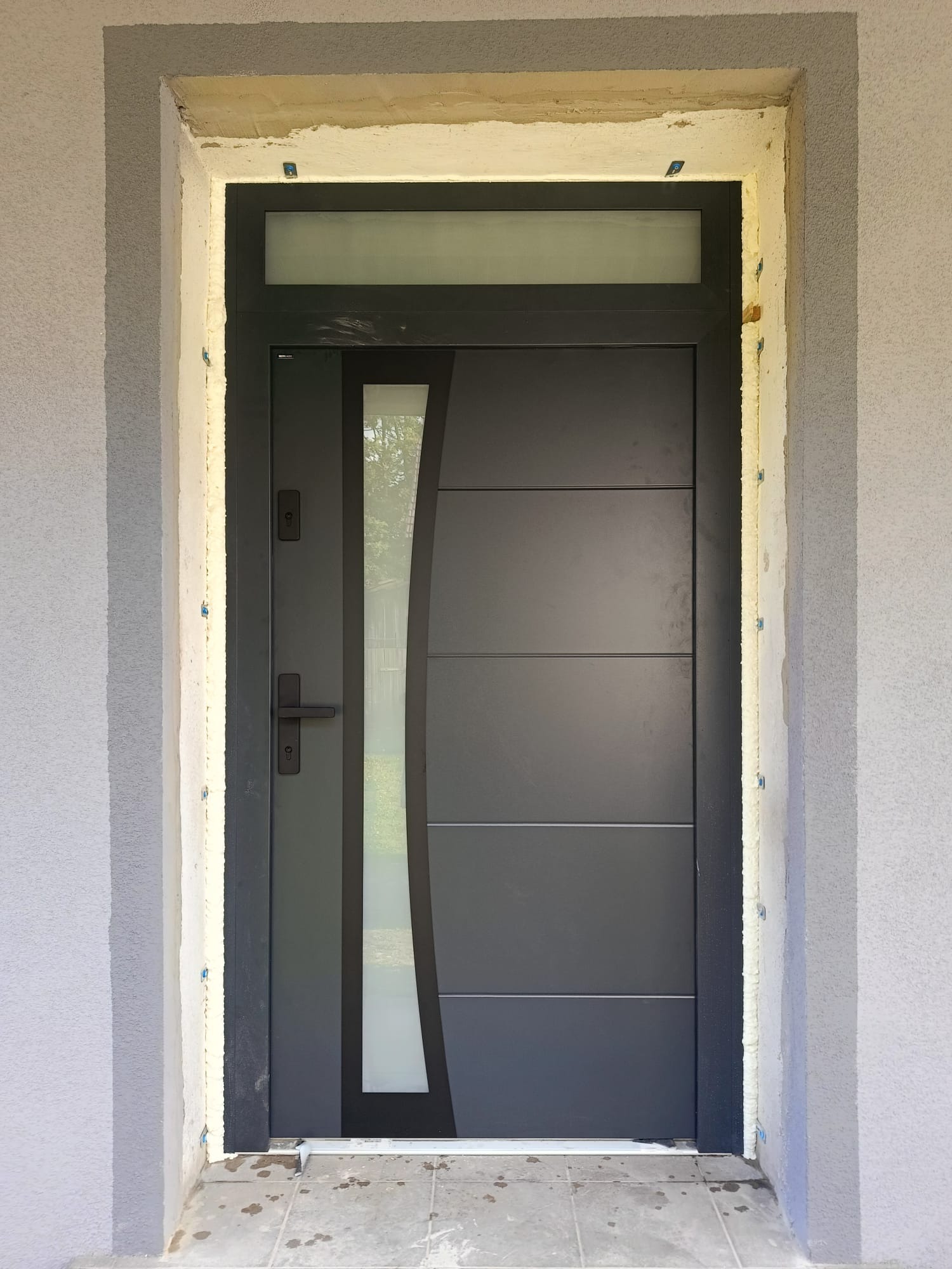 Grafitowe wejściowe z szybą, zamontowane w surowym otworze. Widoczne pianka montażowa i nieotynkowane ściany. Nowoczesny design z poziomymi panelami i pionową szybą.