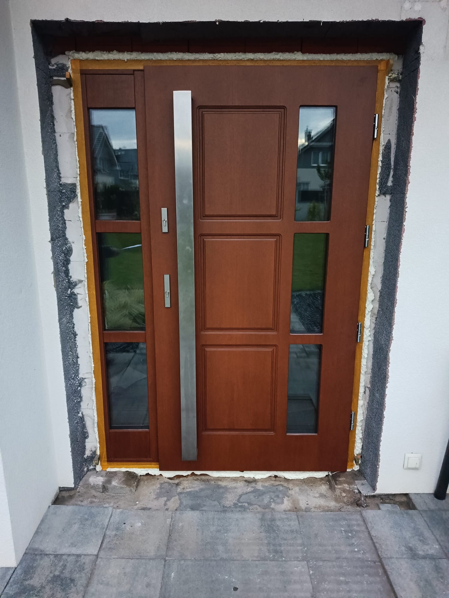 Brązowe drzwi zewnętrzne z szybami i długim, srebrnym uchwytem, w trakcie montażu. Widoczne pianka montażowa i surowy mur. Ujęcie z bliska, z zewnątrz budynku.