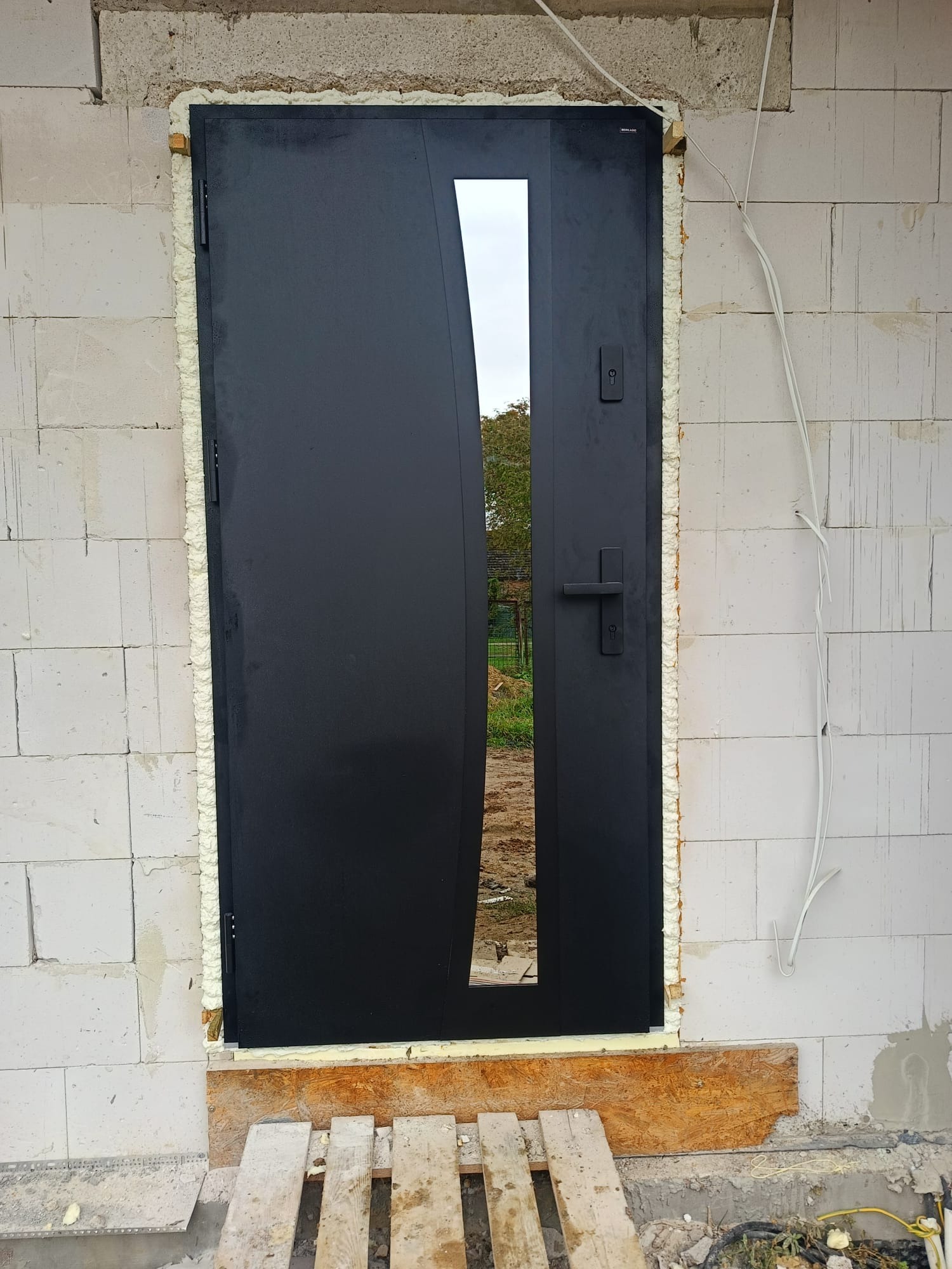Czarne, nowoczesne drzwi z szybą, osadzone w surowym murze z białych bloczków. Widoczna pianka montażowa i drewniane podpory. Ujęcie z bliska, z zewnątrz budynku.