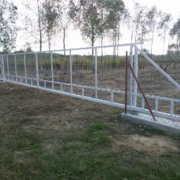 Biała metalowa brama wjazdowa z siatką, częściowo zamontowana, widoczna na tle drzew i trawy, z czerwoną podporą tymczasową.