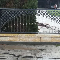 Fragment ogrodzenia kutego z dekoracyjnym wzorem rombów i łuków, osadzonego na murku z piaskowca, na tle zielonego krzewu i podjazdu.