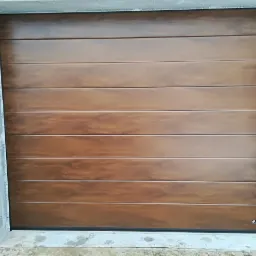 Brązowa, segmentowa brama garażowa z poziomymi panelami, widoczna od frontu, w surowym otworze muru z cegły.