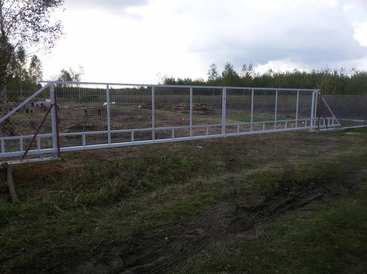 Nowo zainstalowane, przesuwne ogrodzenie panelowe w kolorze białym, widok z zewnątrz na tle drzew i pochmurnego nieba.