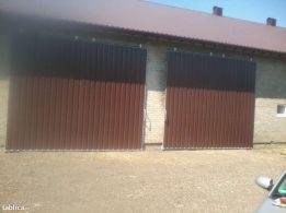 Dwie rozsuwane bramy w stodole, brązowo-czarne, z widocznym mechanizmem otwierania, na tle starego budynku z cegły i dachu z brązowej blachy. Widoczne lekkie zanieczyszczenia na ziemi przed bramami.