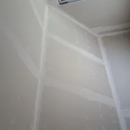 Wnętrze pomieszczenia w trakcie prac wykończeniowych, widoczne płyty gipsowo-kartonowe ze spoinami wypełnionymi masą szpachlową, przygotowane do dalszej obróbki, z widocznym fragmentem sufitu...