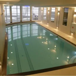 Wnętrze krytego basenu z jasnymi płytkami, dekoracyjnymi pasami na ścianach i dużymi oknami w tle, oświetlonego punktowymi lampami.