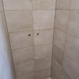 Wykończona kabina prysznicowa z beżowymi, matowymi płytkami, widoczne dwa punkty podłączenia baterii i liniowy odpływ podłogowy.