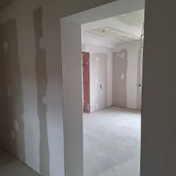 Wnętrze w trakcie prac wykończeniowych: ściany z płyt kartonowo-gipsowych z widocznymi łączeniami wymagającymi szpachlowania, surowy betonowy podkład, widoczne instalacje elektryczne na suficie.