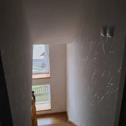 Klatka schodowa z drewnianymi schodami, ściany pokryte tapetą z efektem marmuru, widok na okno z parapetem i fragmentem ogrodu.