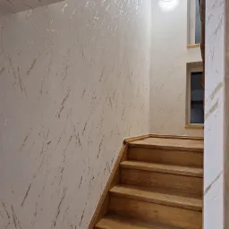 Klatka schodowa z drewnianymi schodami i ścianą pokrytą dekoracyjną, jasną strukturą przypominającą marmurkowe żyłki. Oświetlenie punktowe nad schodami.