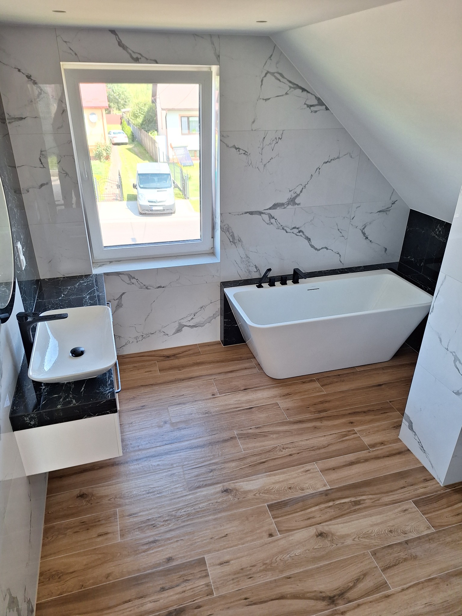 Wykończona łazienka na poddaszu z drewnopodobnymi płytkami podłogowymi, białą wanną i umywalką nablatową na czarnym blacie z efektem marmuru, ściany wyłożone płytkami imitującymi biały marmur...