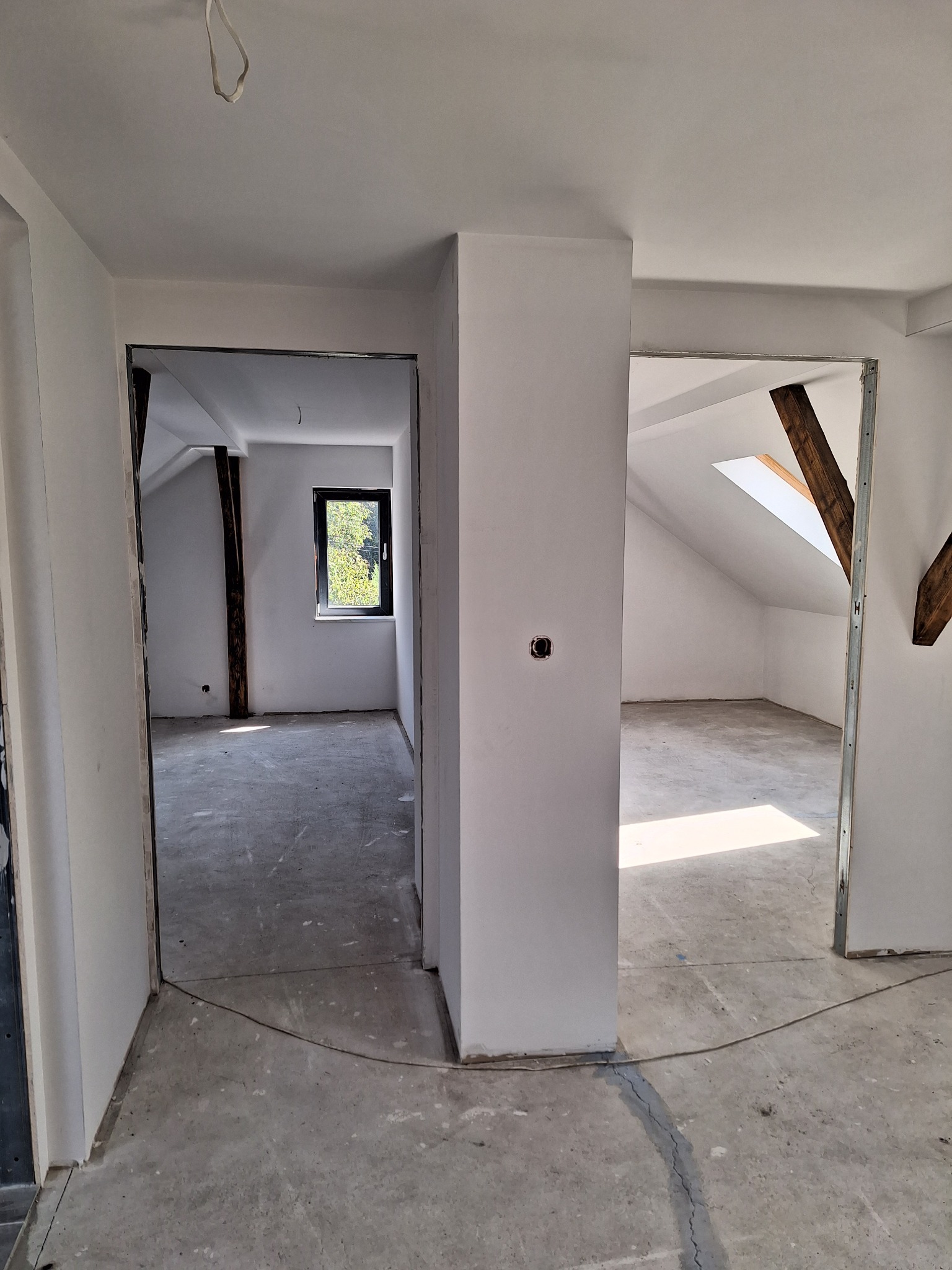 Wnętrze poddasza po wyrównaniu ścian, widoczne dwa otwory drzwiowe z metalowymi profilami, surowe ściany przygotowane do dalszych prac wykończeniowych, okno dachowe w jednym z pomieszczeń.