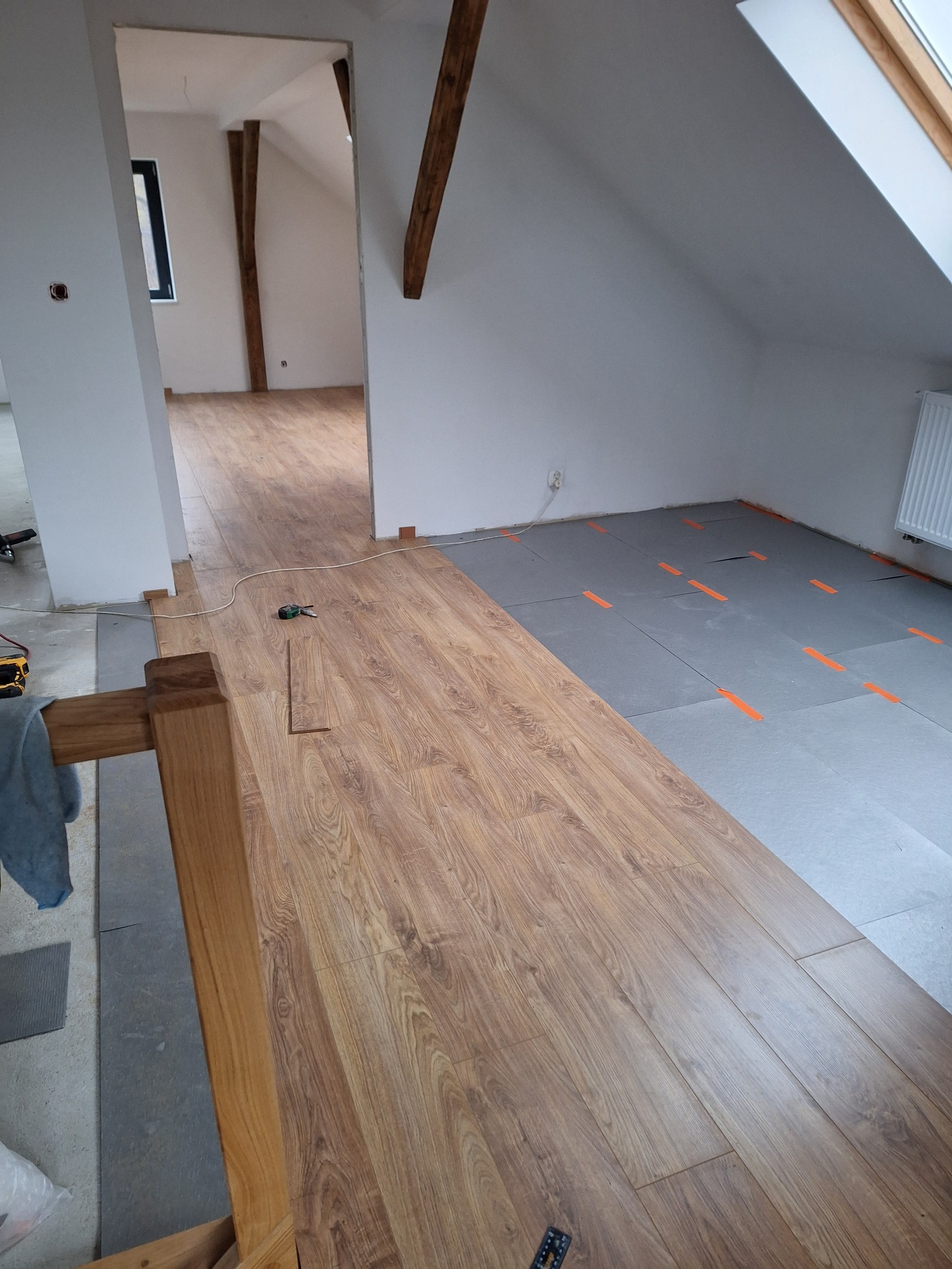 Montaż paneli podłogowych w pokoju na podkładzie izolacyjnym, widoczne narzędzia pomiarowe i fragment drewnianej konstrukcji schodów.