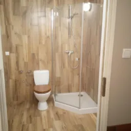 Wyremontowana łazienka z drewnopodobnymi płytkami na ścianach i podłodze, białą toaletą z drewnianą deską sedesową oraz narożną kabiną prysznicową z chromowaną armaturą.