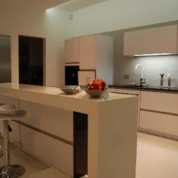 Nowoczesna kuchnia z białymi szafkami, czarnym blatem, wyspą z białego materiału i hokerami. Na wyspie miski z owocami. Oświetlenie podszafkowe LED.