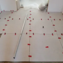 Układanie jasnych, kwadratowych płytek podłogowych z użyciem poziomicy i czerwonych klipsów dystansowych, folia ochronna na ścianach w tle.