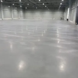 Gładka, szara posadzka w hali przemysłowej z metalowymi ścianami i oświetleniem sufitowym, widoczne delikatne refleksy świetlne na powierzchni.