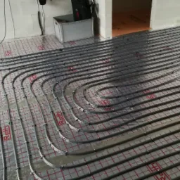 Szare rury ogrzewania podłogowego ułożone w pętle na folii z czerwoną kratką, widoczne narzędzia i skrzynka w tle.