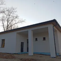 Parterowy dom z płaskim dachem, świeżo otynkowany na biało, z widocznymi kolumnami wejściowymi i czarną stolarką okienną, na tle jesiennego krajobrazu z drzewami bez liści i latającymi ptakami.