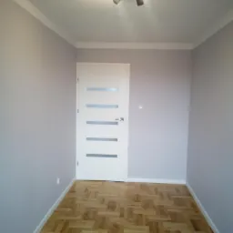 Pokój po remoncie: świeżo pomalowane ściany w odcieniach szarości, białe listwy przypodłogowe i sufitowe, parkiet ułożony w jodełkę, białe drzwi z poziomymi przeszkleniami i czarna lampa sufitowa...