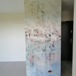 Ściana działowa z nadrukiem przedstawiającym flamingi w wodzie, widoczne gniazdko elektryczne na dole, szara podłoga.