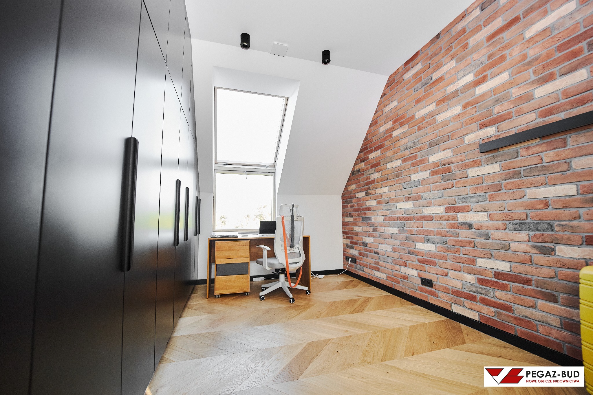 Pokój biurowy na poddaszu z ceglaną ścianą, drewnianą podłogą w jodełkę, biurkiem z laptopem pod oknem dachowym i dużą, czarną szafą wnękową.