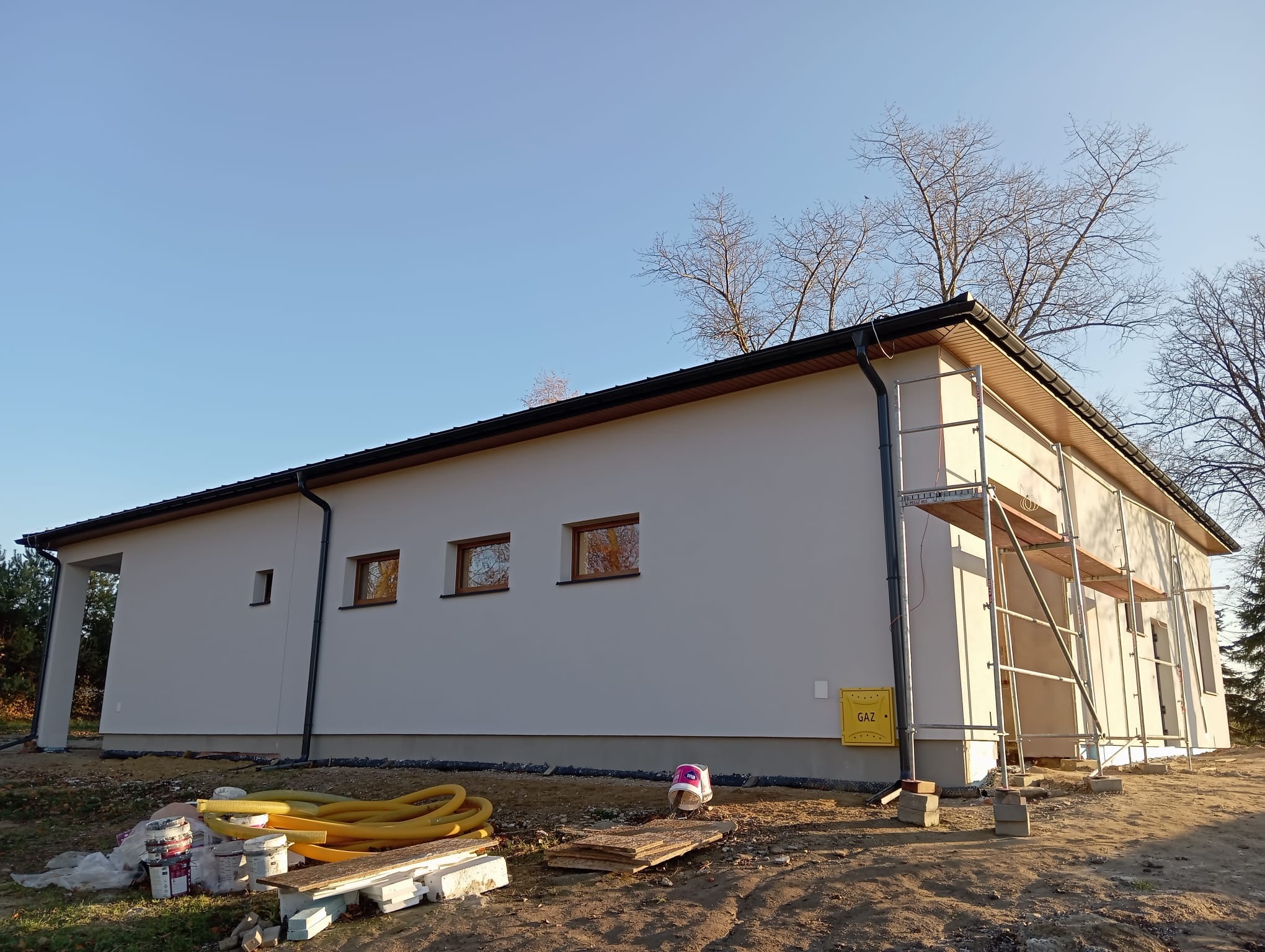 Nowo wybudowany dom jednorodzinny z jasną elewacją, widoczne rusztowanie, okna z brązowymi ramami, żółte rury wentylacyjne i puszki farb na ziemi.